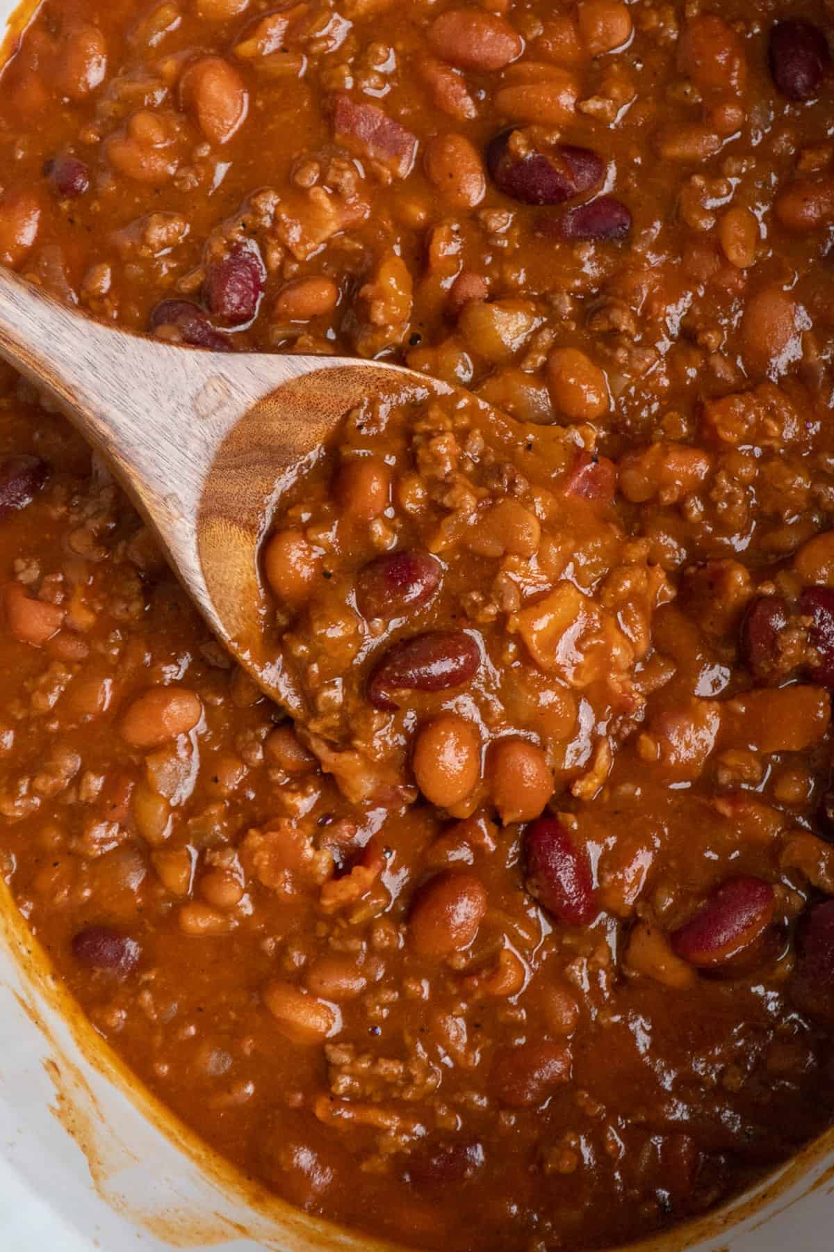 Cowboy beans in a white slow cooker with a wooden spoon.