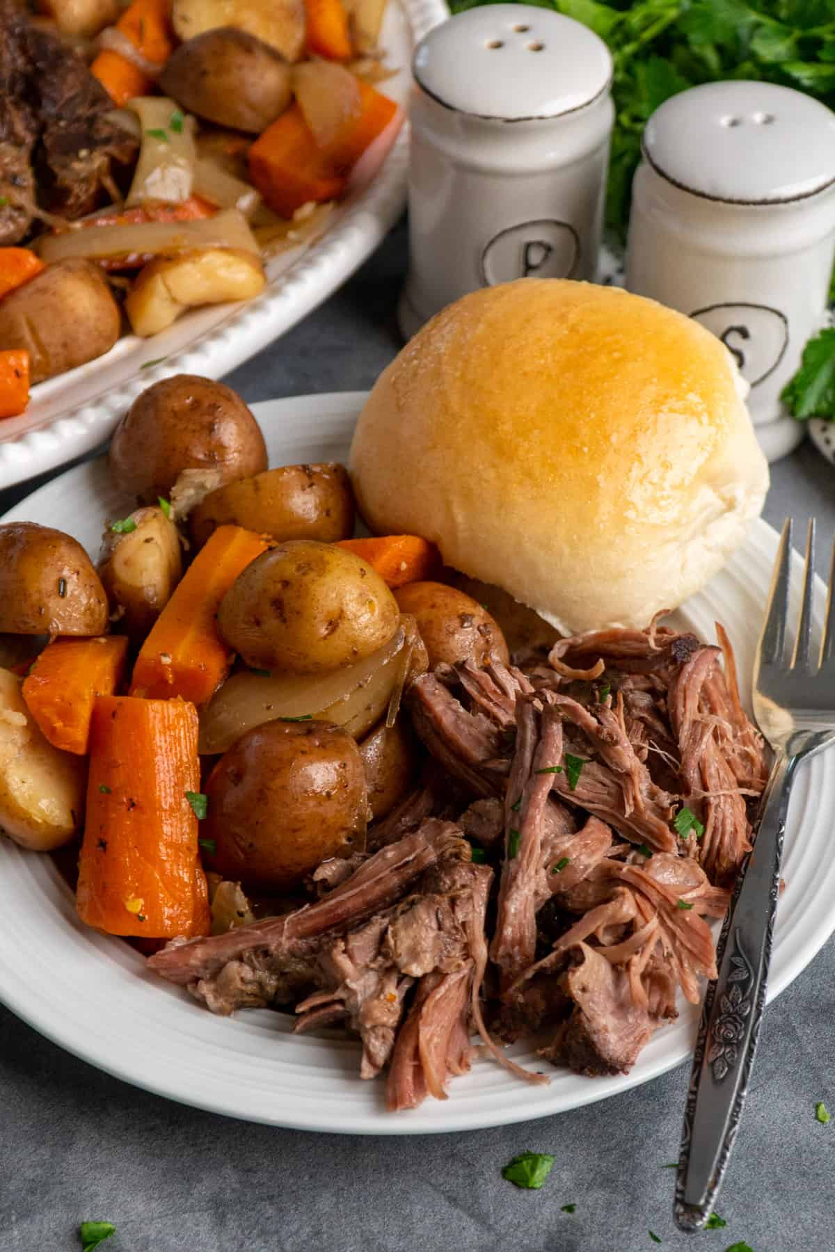 Shredded beef roast on a plate with a roll and vegetables.
