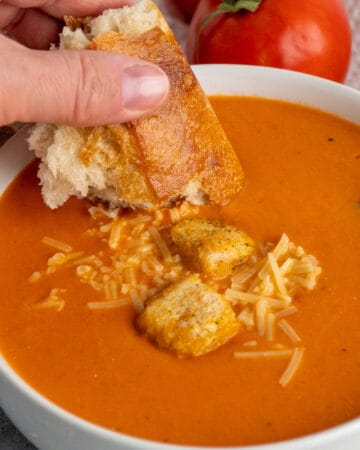 A hand dipping a piece of bread into creamy tomato soup.
