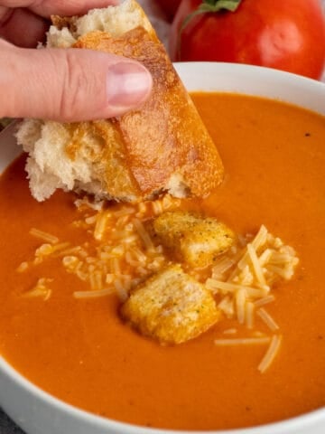 A hand dipping a piece of bread into creamy tomato soup.