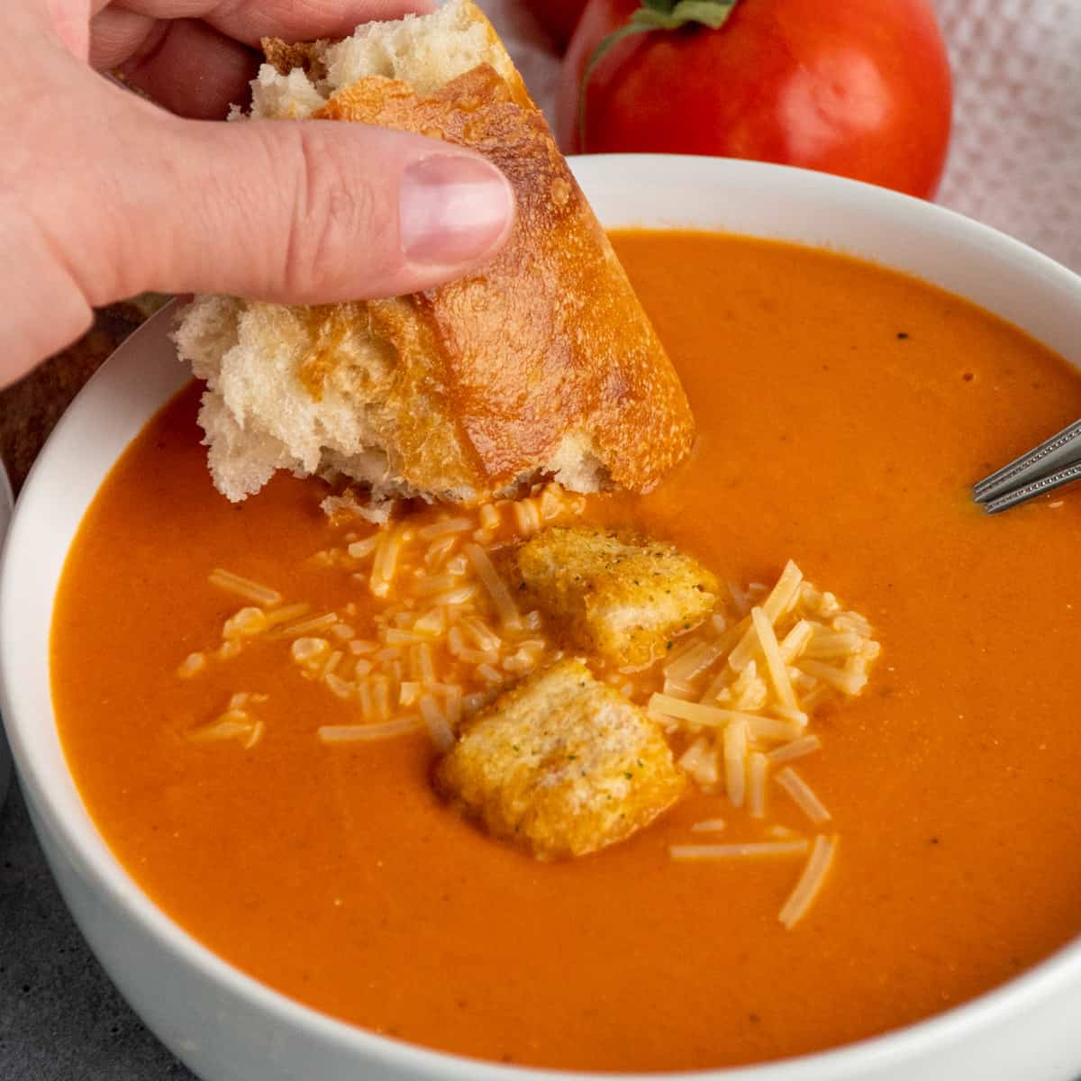 A hand dipping a piece of bread into a bowl of tomato soup.