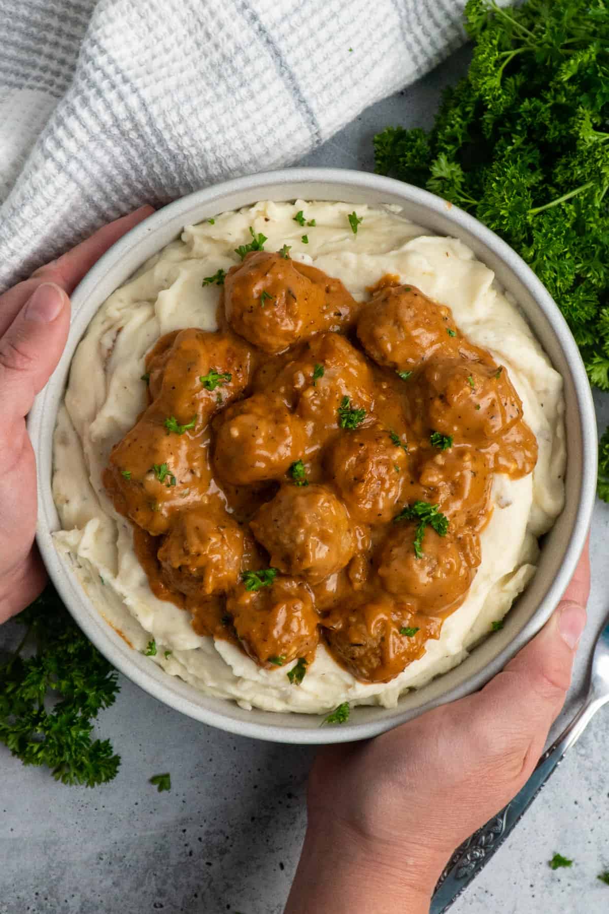 Hands holding a bowl of meatballs and gravy over mashed potatoes.