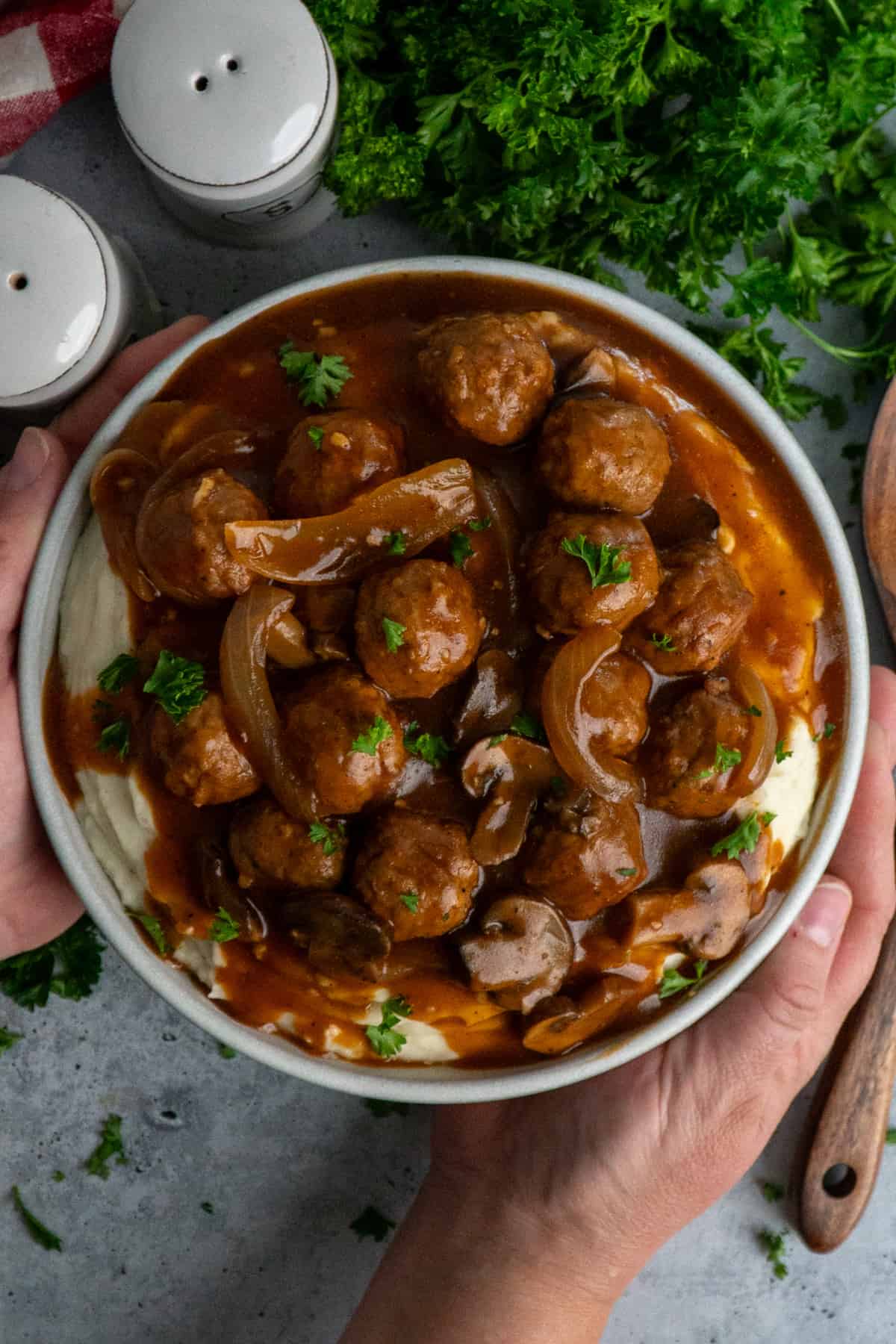 Hands holding a bowl of salisbury steak meatballs over mashed potatoes.