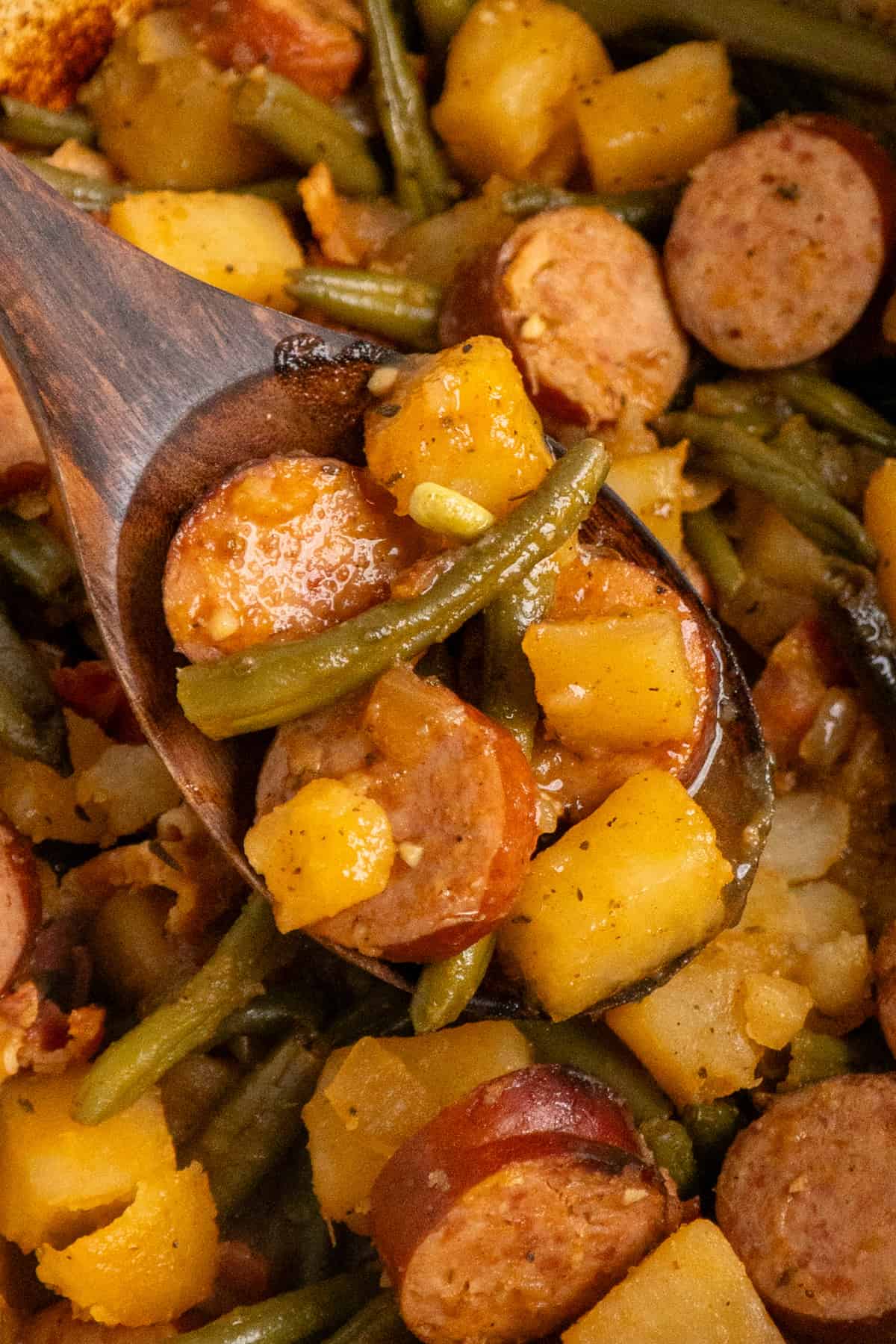 A wooden spoon holding a scoop of sausage potatoes and green beans.
