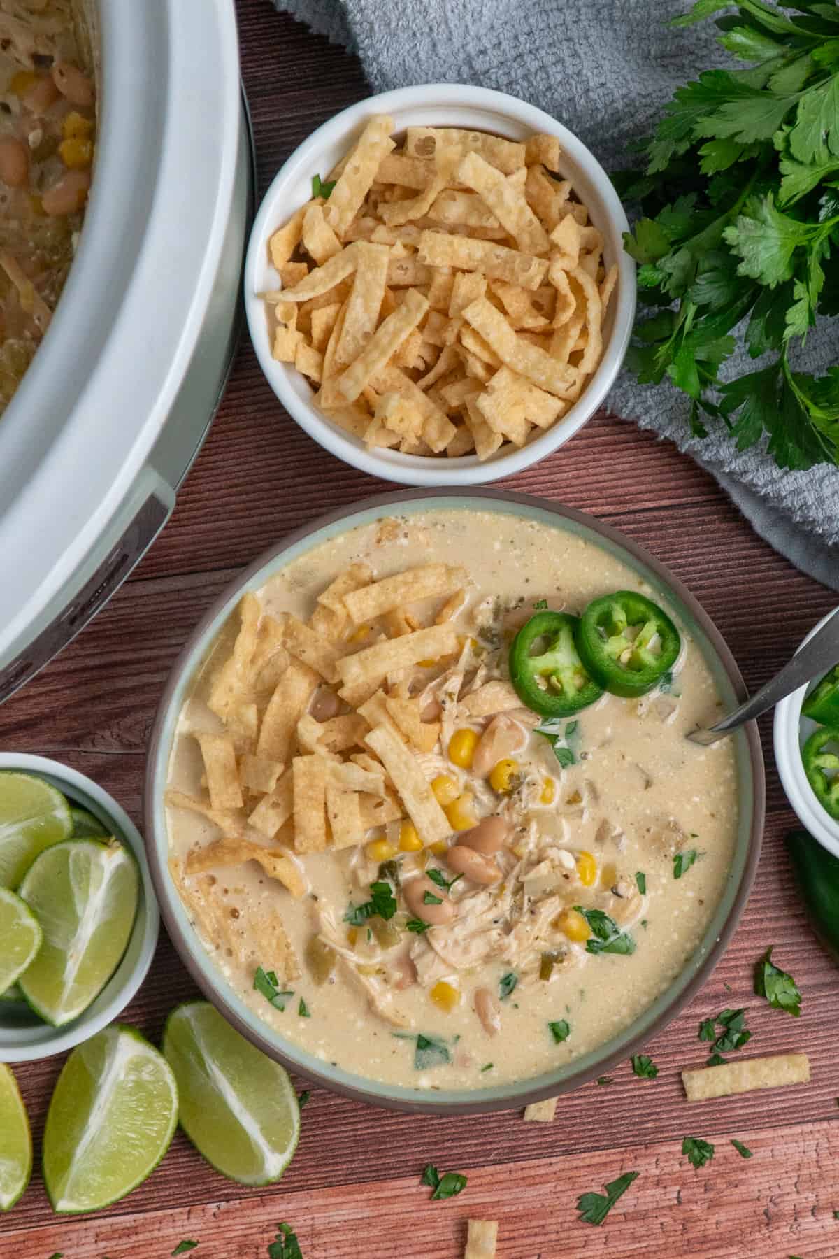 White chicken chili in a bowl and in a slow cooker on the table with parsley and a bowl of tortilla strips.