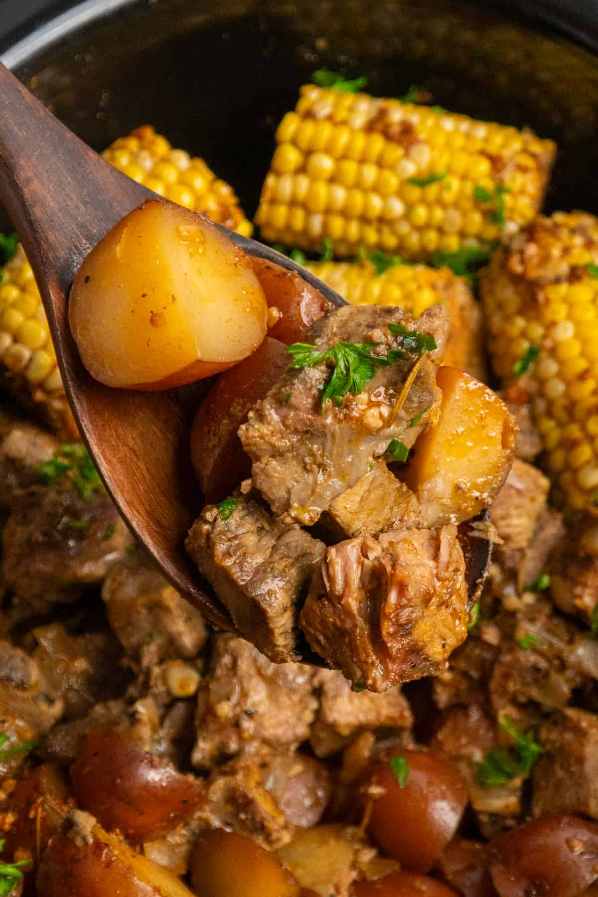 A wooden spoon holding a scoop of steak and potatoes over a slow cooker.