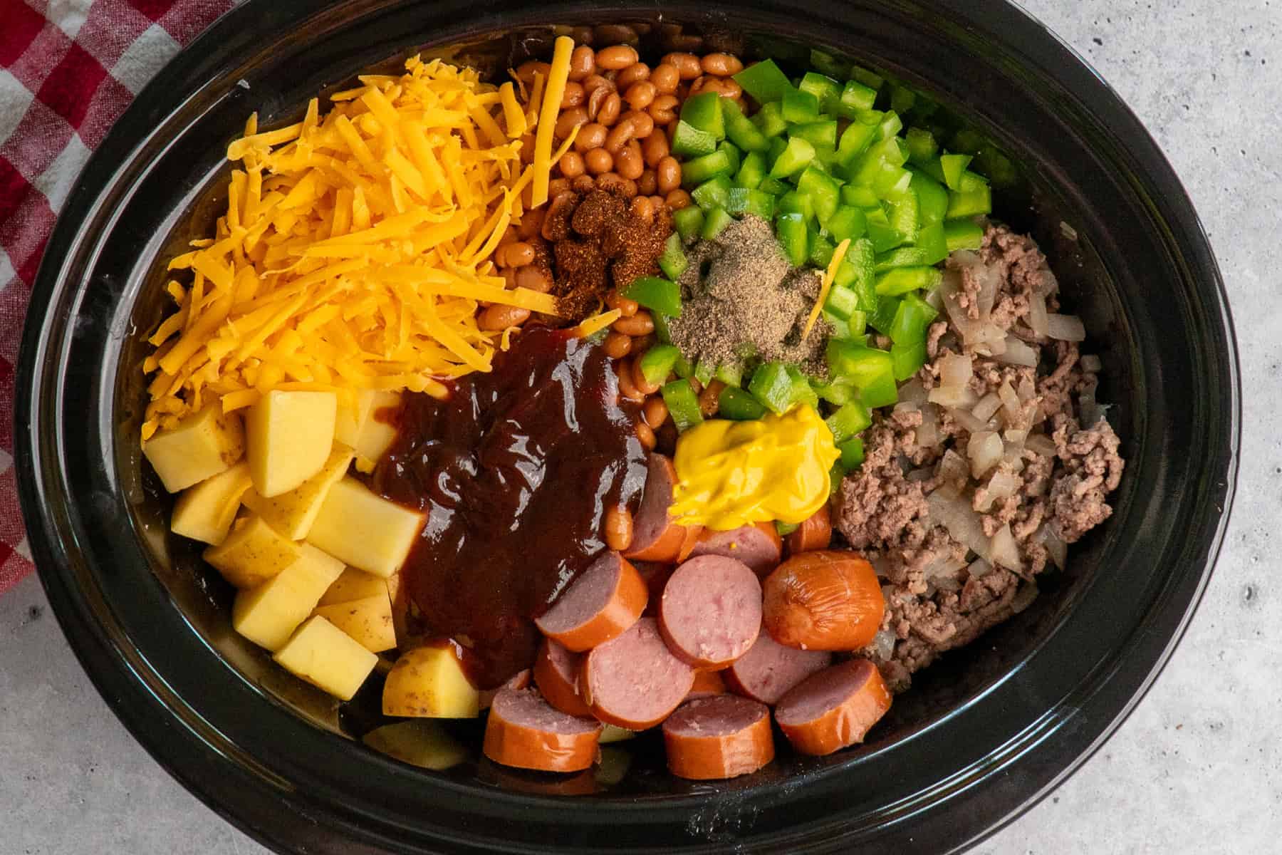 Overhead look at cheese, potatoes, spices, bbq sauce, sausage, ground beef, onions and green peppers in a slow cooker.