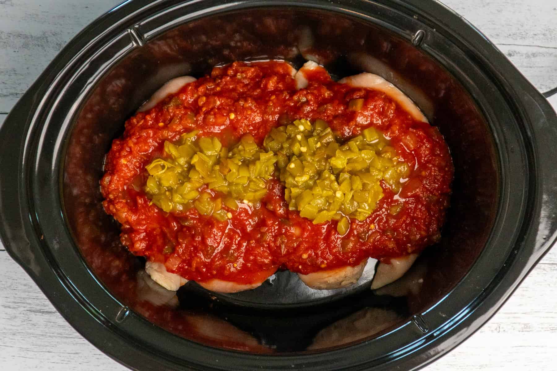 Overhead look at chicken breasts, salsa, and diced green chilies in a crock pot.