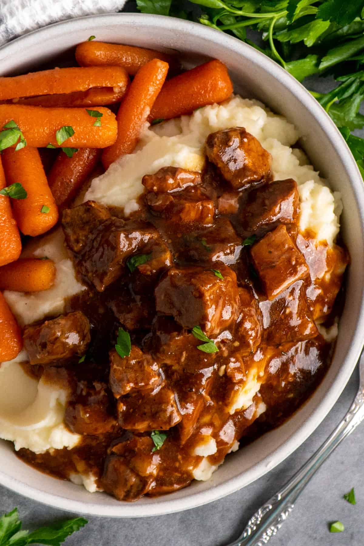 Beef tips and gravy over mashed potatoes with a side of carrots all garnished with chopped parsley.