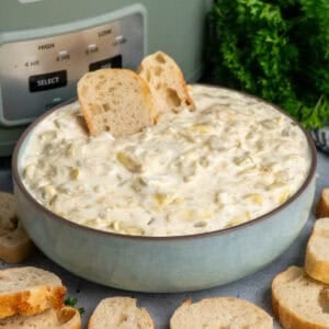 Artichoke dip in a blue bowl with baguette slices with a crock pot in the background.