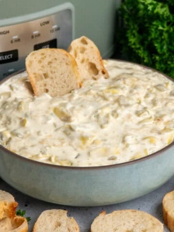 Artichoke dip in a blue bowl with baguette slices with a crock pot in the background.