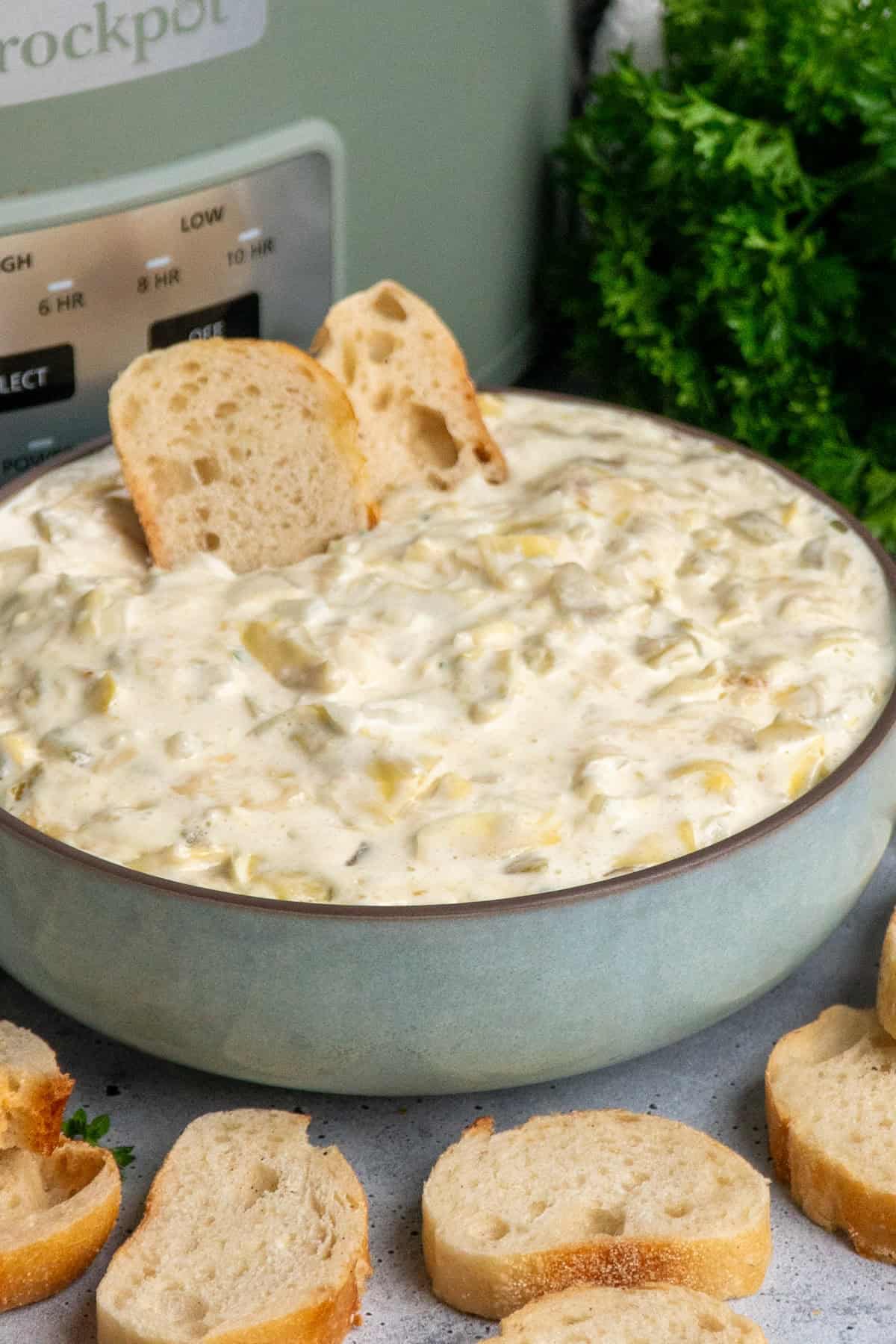 Artichoke dip in a blue bowl with baguette slices with a crock pot in the background.