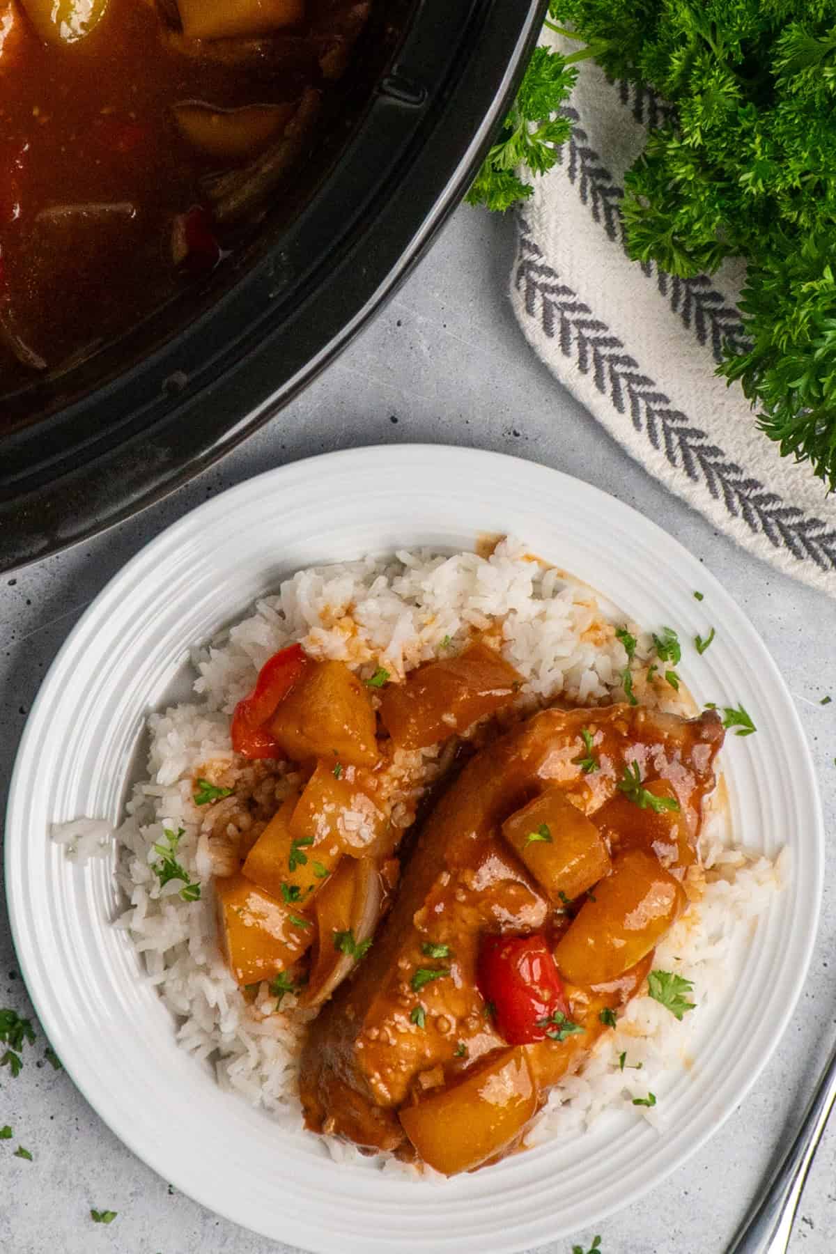 A Hawaiian pork chop on a plate of rice with a crock pot in the background.