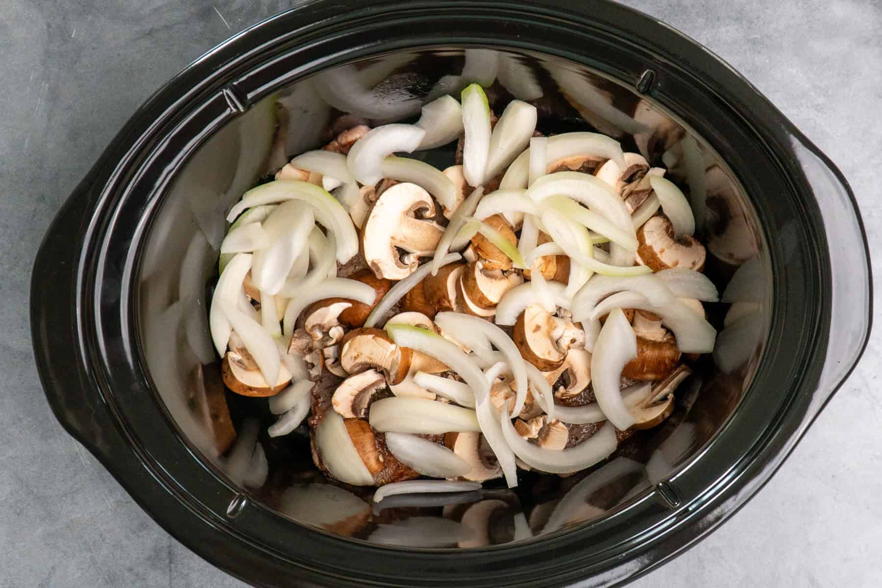 Mushrooms and onions covering a steak in a crock pot.