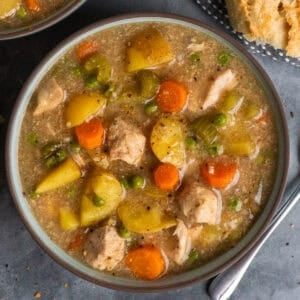 A bowl of slow cooker chicken stew on a table with a spoon, bread, and more stew around the edges of the bowl.