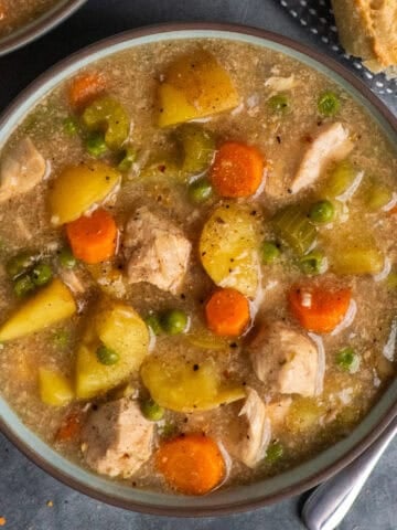 A bowl of slow cooker chicken stew on a table with a spoon, bread, and more stew around the edges of the bowl.