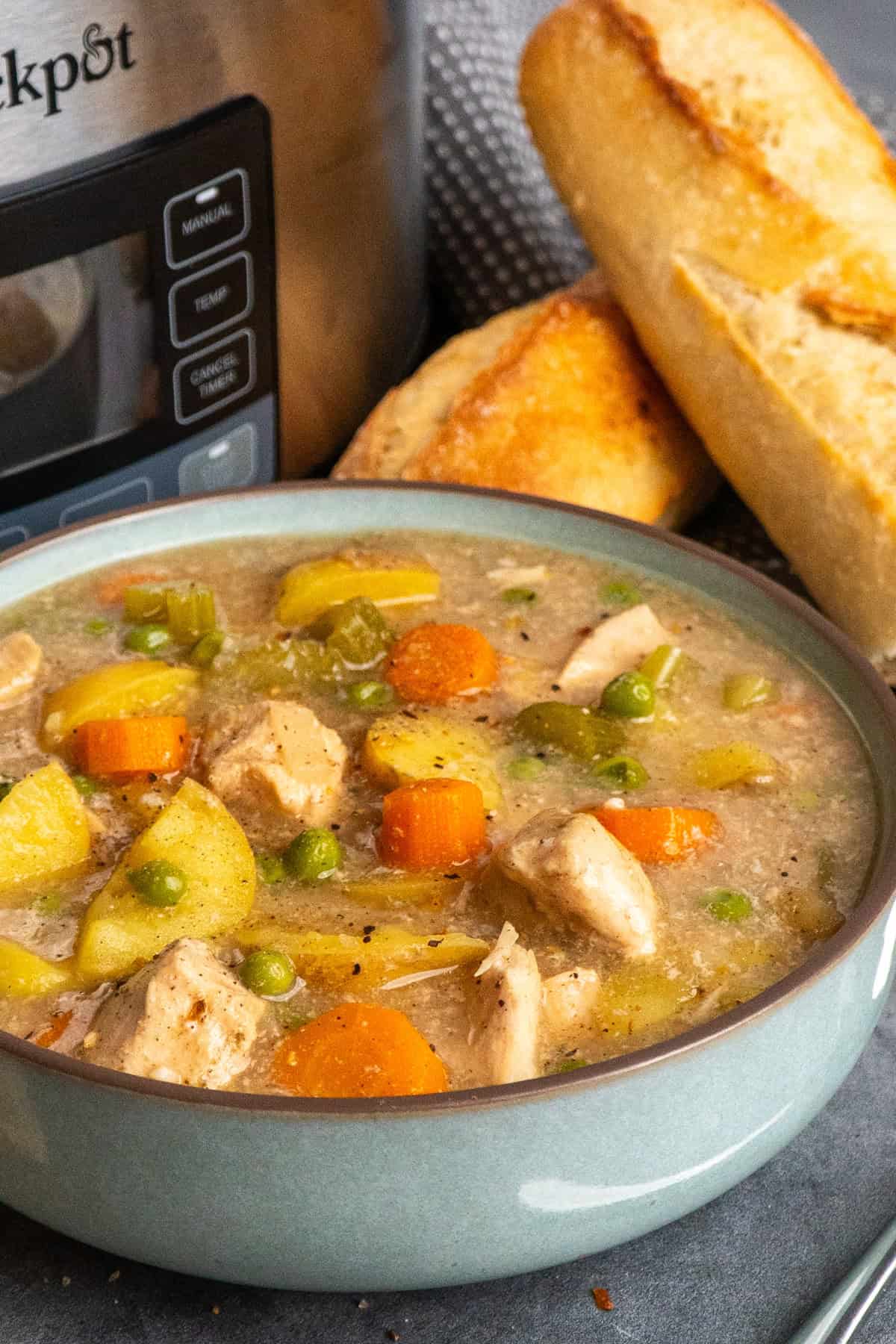 A bowl of chicken stew on the table in front of a slow cooker with a loaf of French bread to the side.