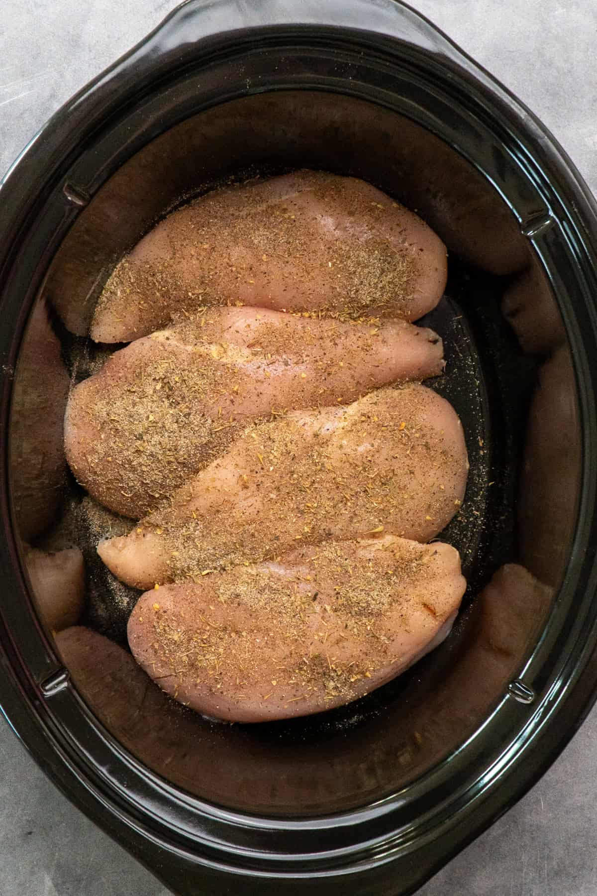 Four chicken breasts in a slow cooker with seasoning on them.