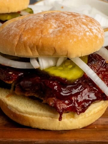 BBQ rib sandwich on a wooden cutting board with pickles and onions.