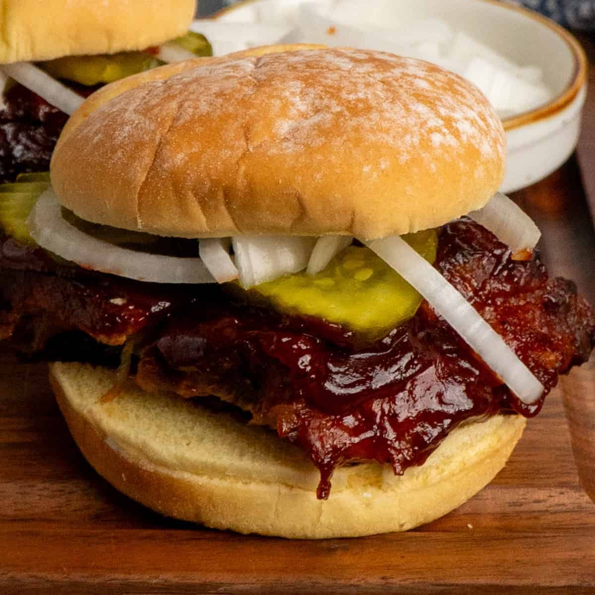 BBQ rib sandwich on a wooden cutting board with pickles and onions.