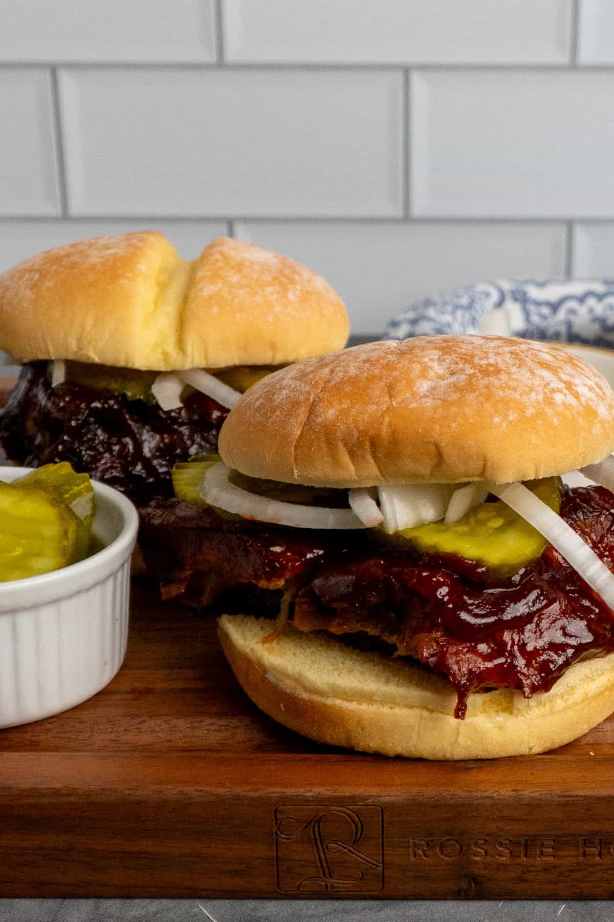 Two bbq rib sandwiches on a cutting board with a bowl of pickles to the side.