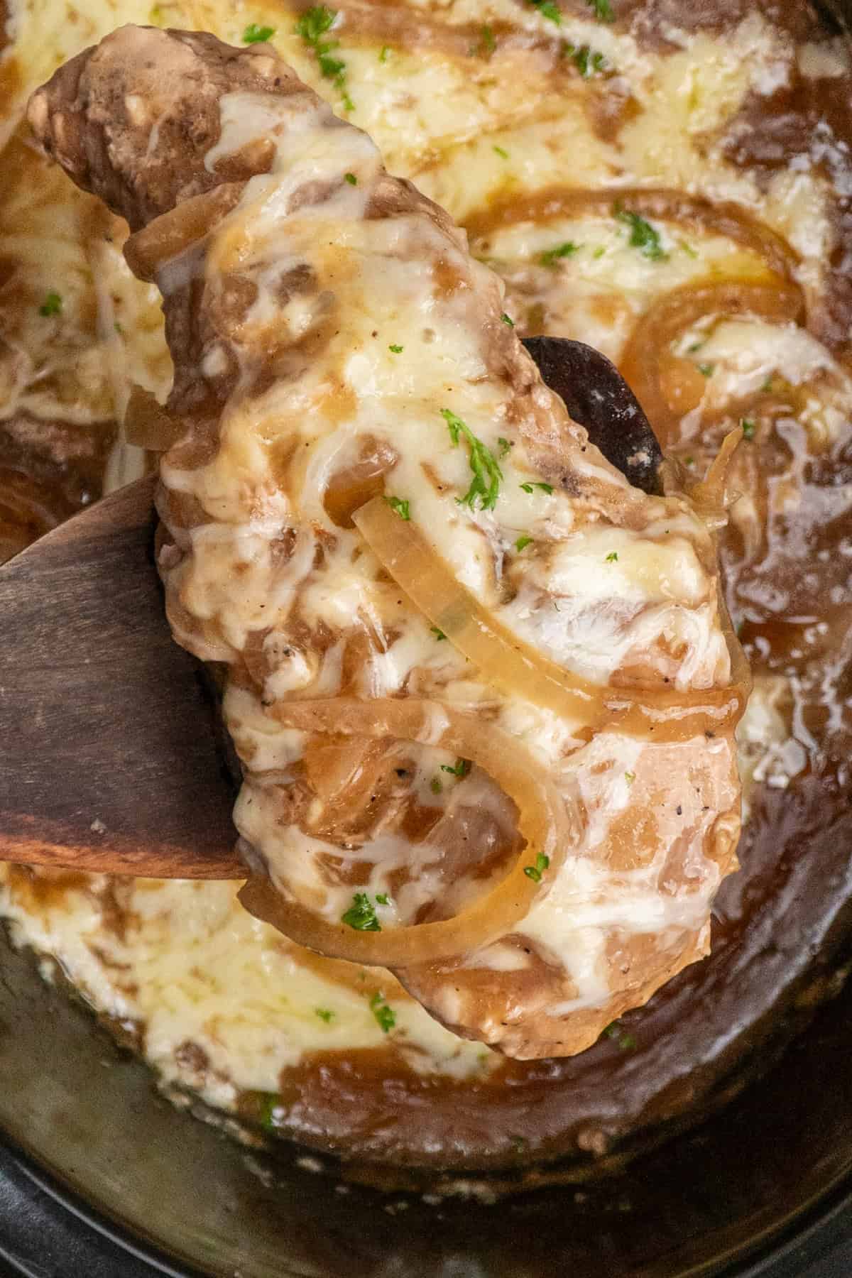 A wooden spatula holding a French onion pork chop over a slow cooker.