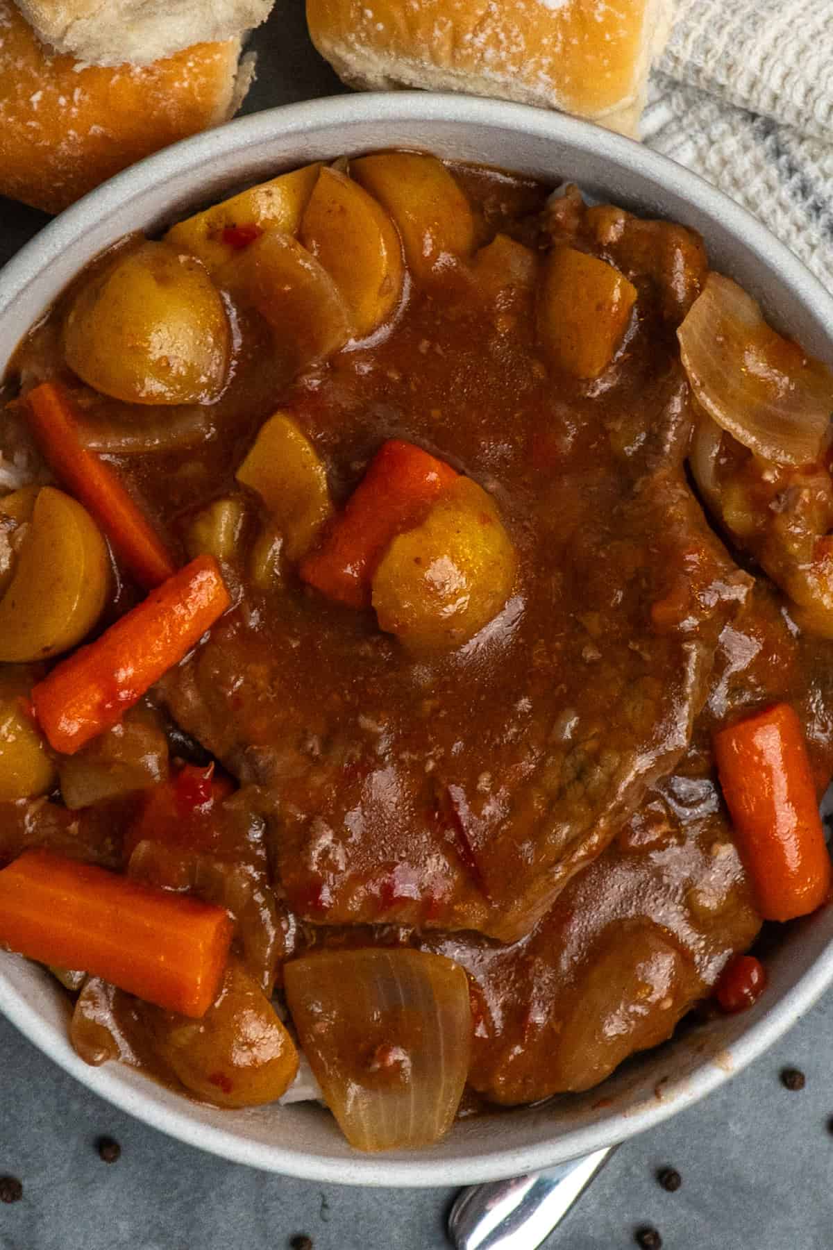 A bowl of crockpot Swiss steak on the table with some rolls and a white tea towel.