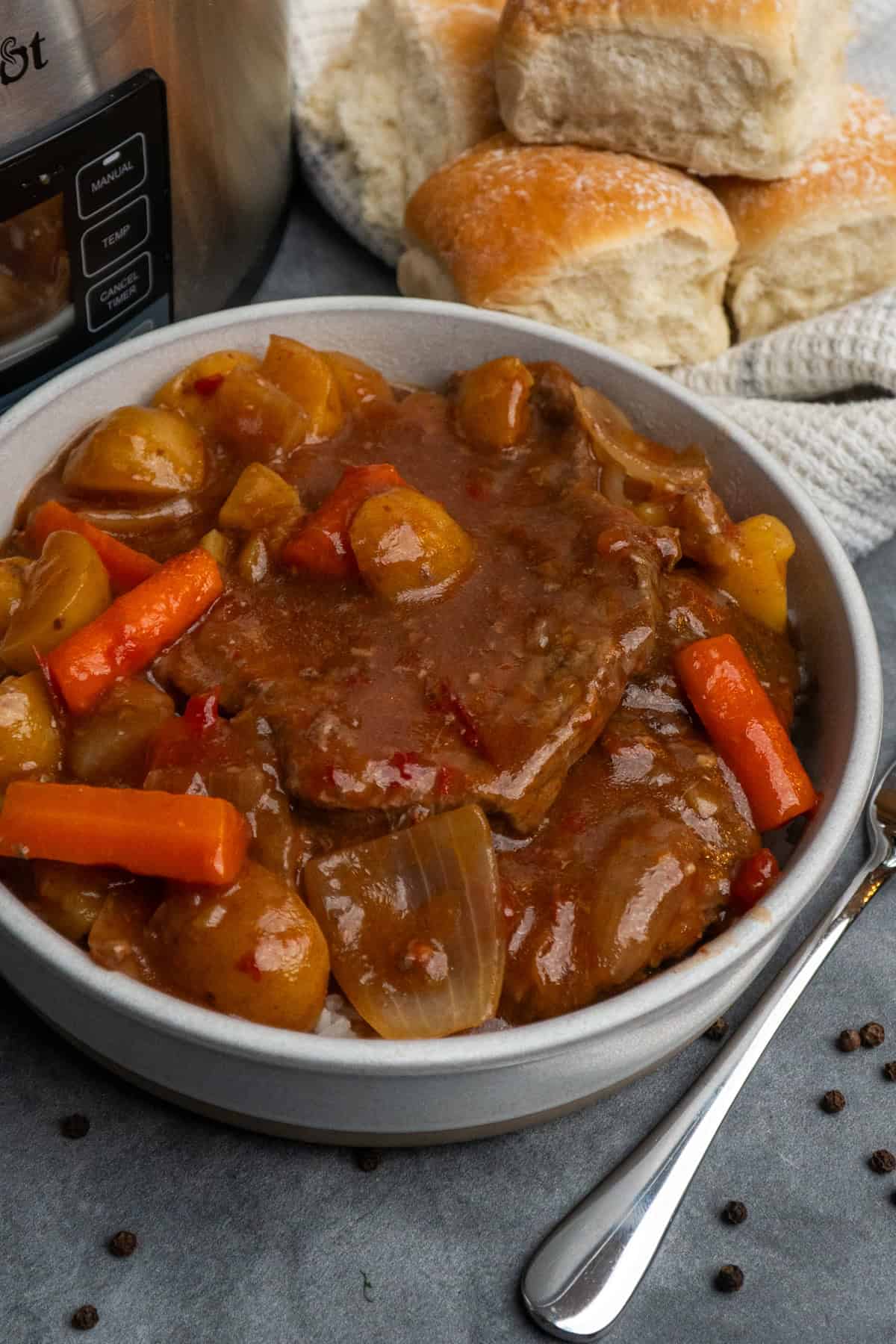 A bowl of crockpot Swiss steak in a bowl with a basket of rolls in the background. 