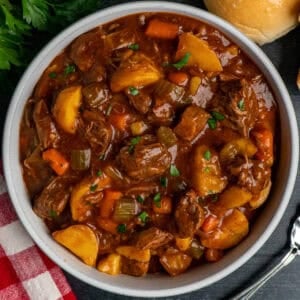 Slow cooker beef stew in a white bowl with parsley and a roll in the background.