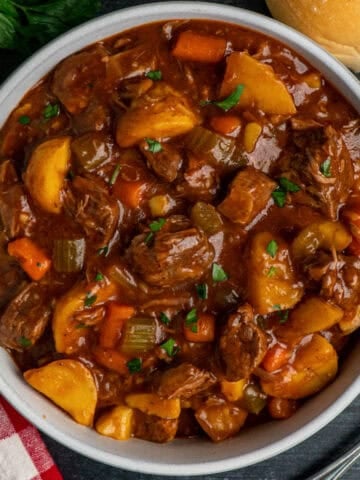 Slow cooker beef stew in a white bowl with parsley and a roll in the background.