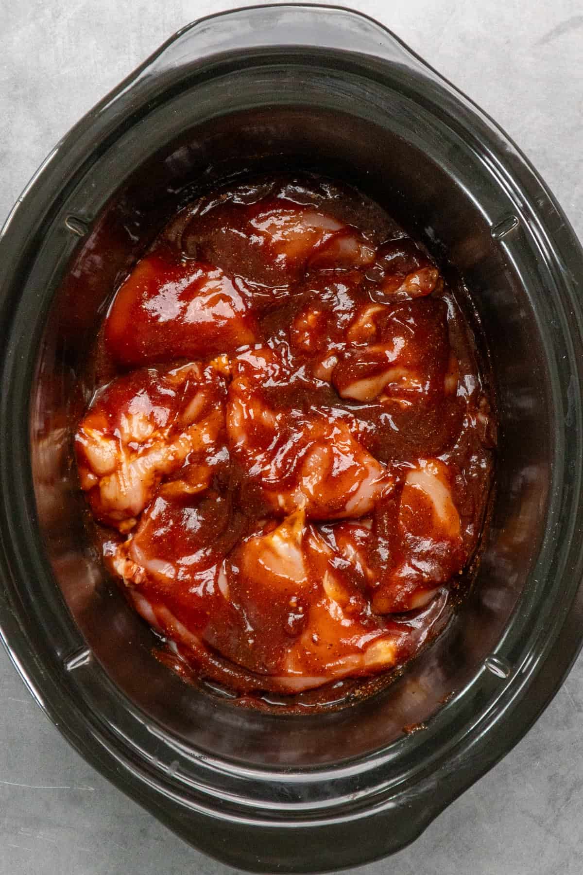 Chicken thighs covered in BBQ sauce and spice in a black slow cooker.