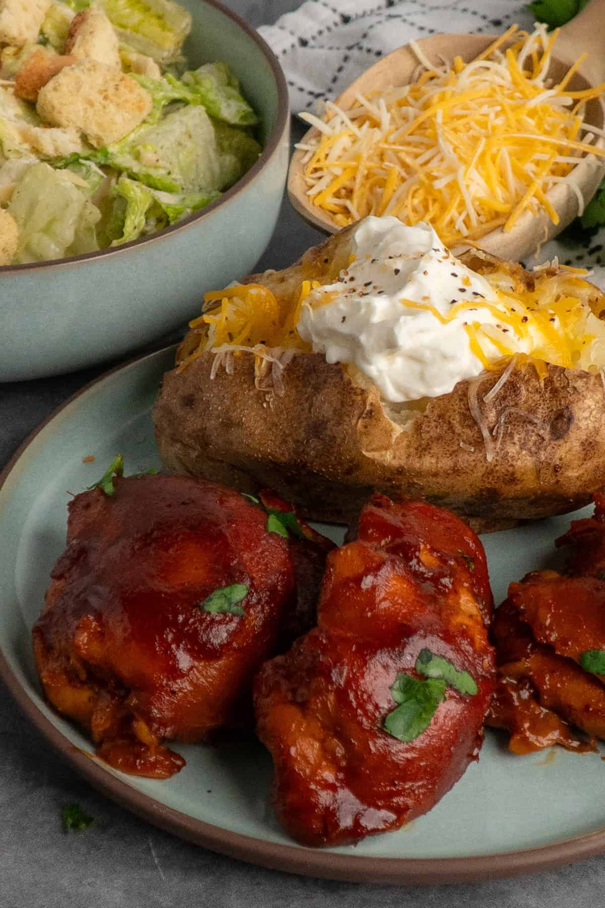 Three BBQ chicken thighs on a plate with a baked potato and salad in the background.