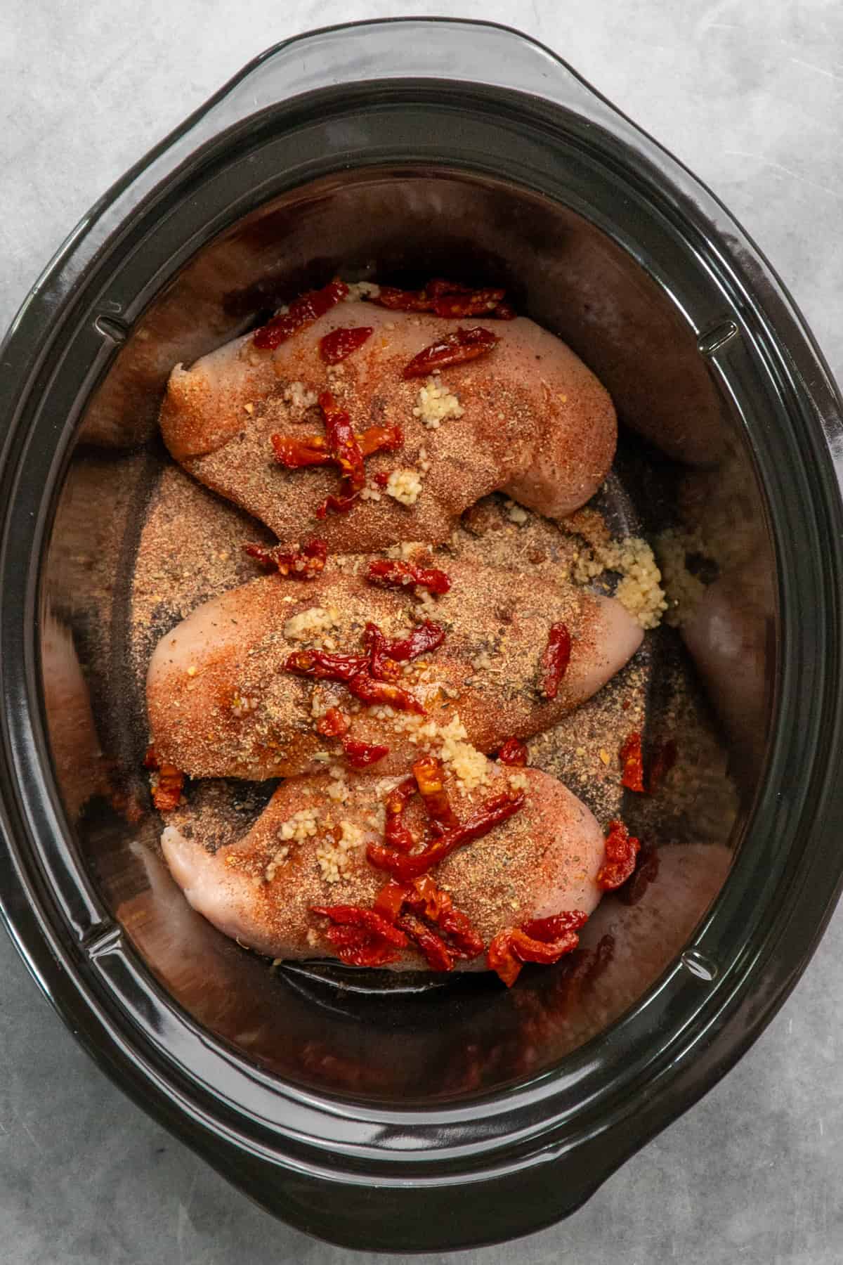 Chicken in a crock pot with seasonings, garlic and sun dried tomatoes on top.