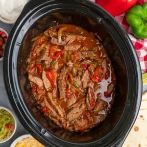 Steak fajitas in a slow cooker with tortillas, cheese, guacamole and sour cream in the background.