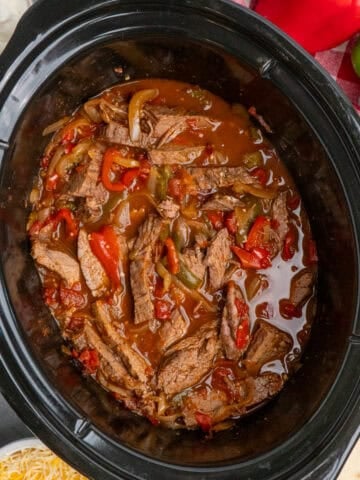 Steak fajitas in a slow cooker with tortillas, cheese, guacamole and sour cream in the background.