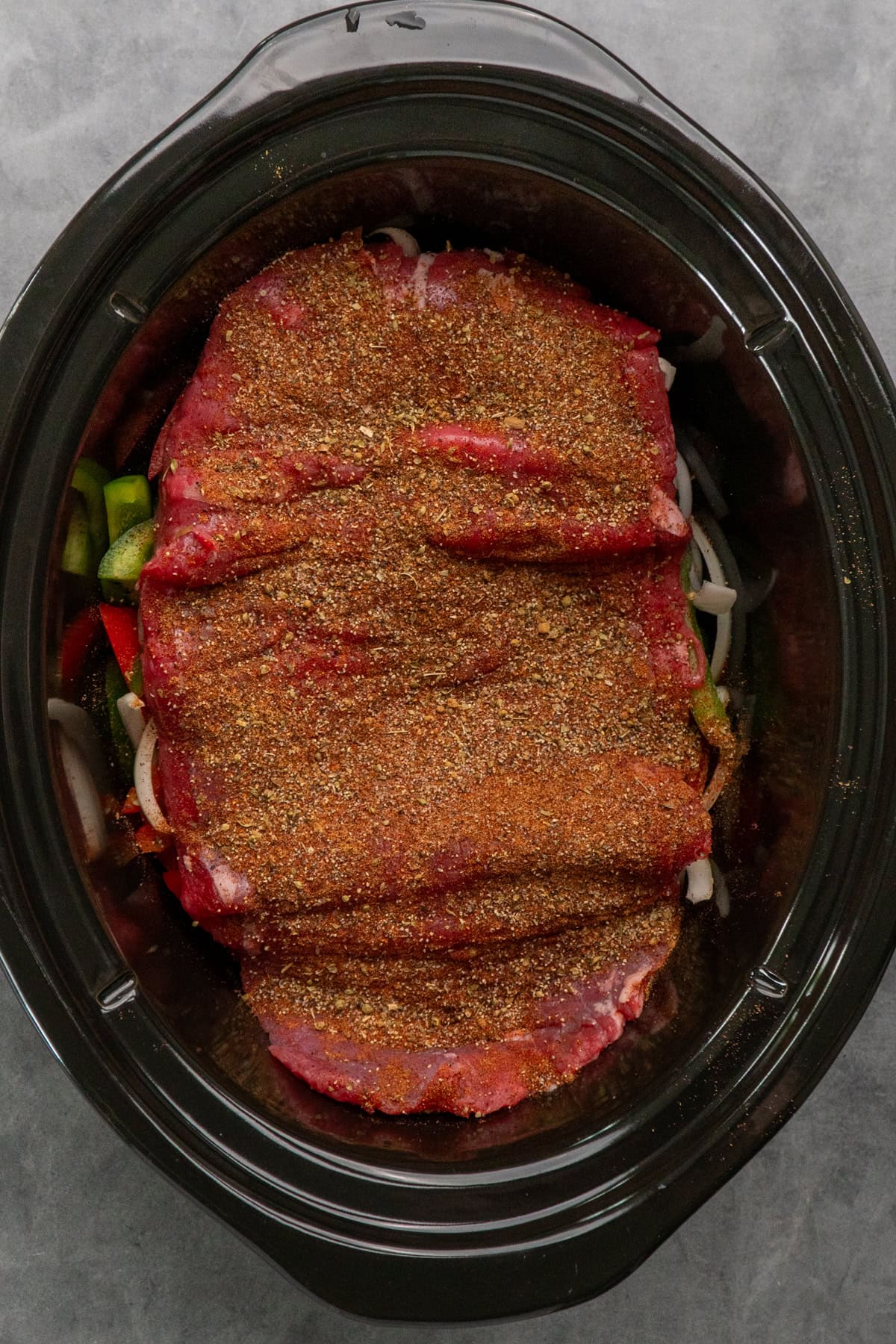 Seasoned flank steak on top of onions and bell peppers in a slow cooker.