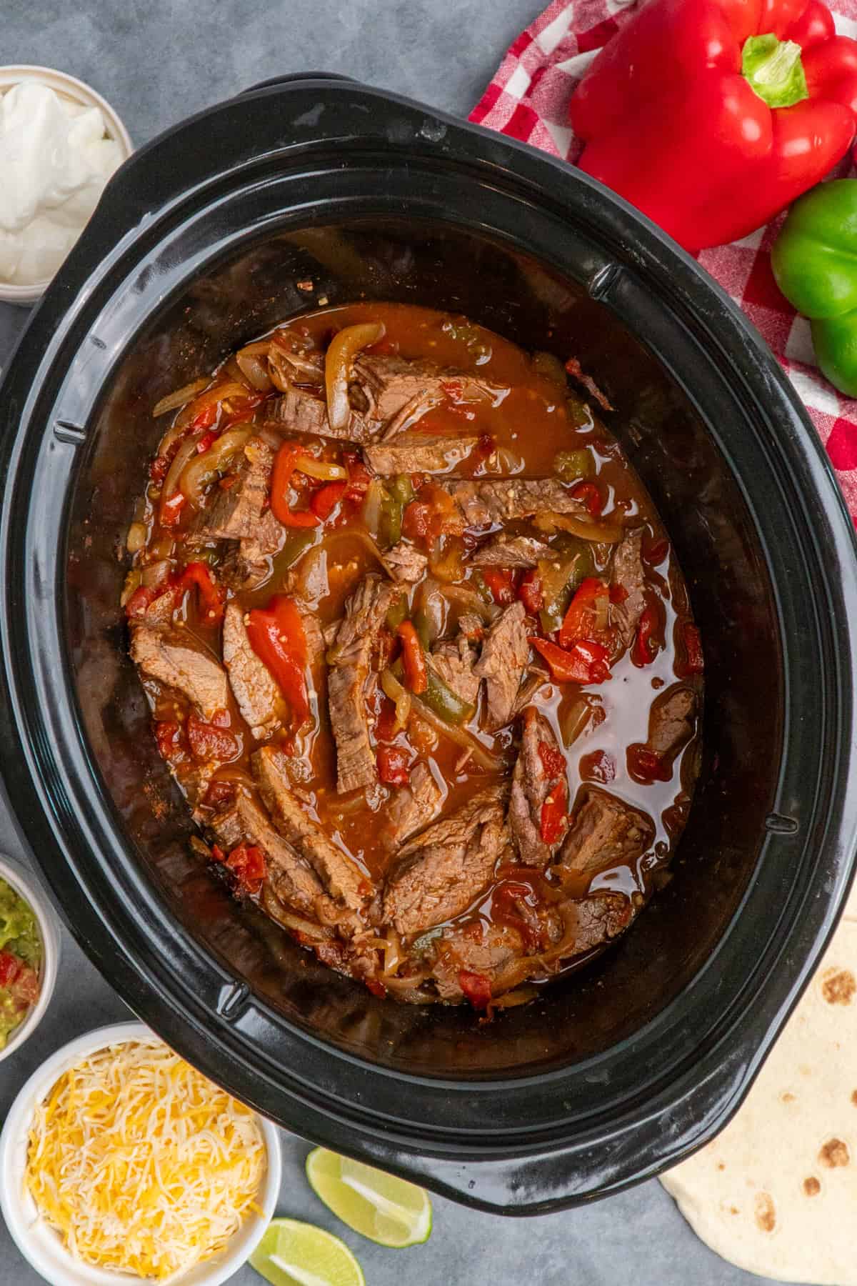 Steak fajitas in a slow cooker with tortillas, cheese, guacamole and sour cream in the background.