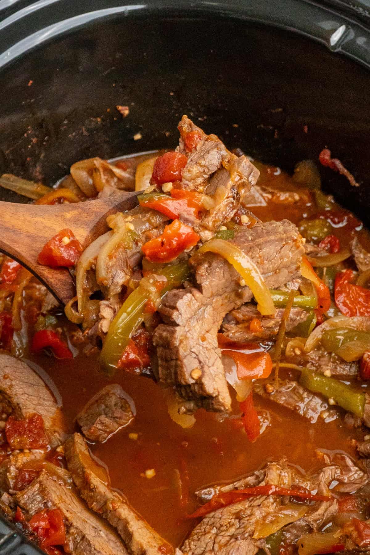 A wooden spoon holding a scoop of steak fajitas over a slow cooker.