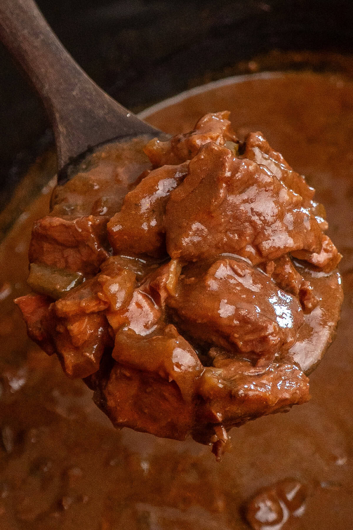 A wooden spoon holding a scoop of steak and gravy over a crock pot.