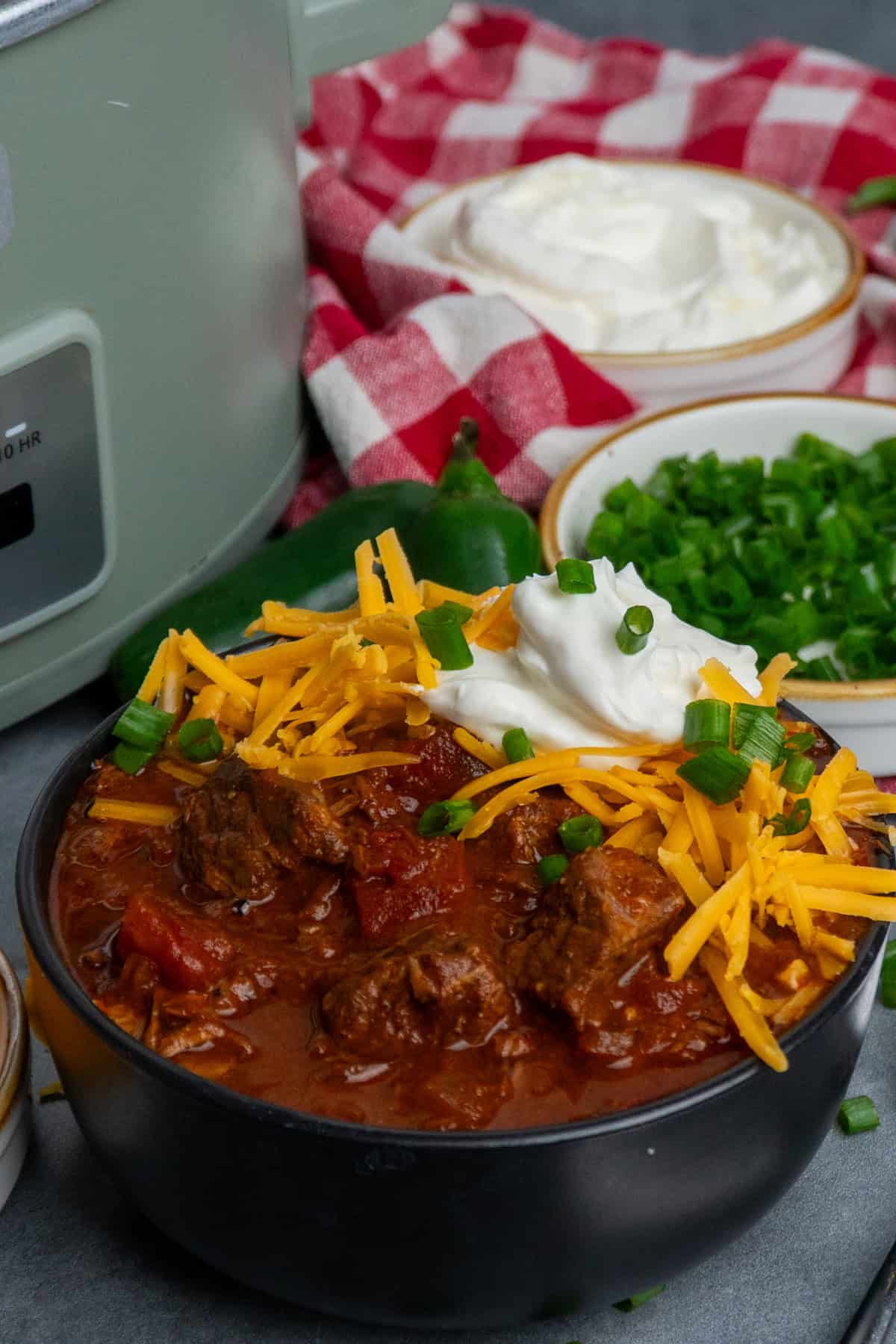 A bowl of Texas chili topped with cheese and sour cream with a slow cooker in the back.