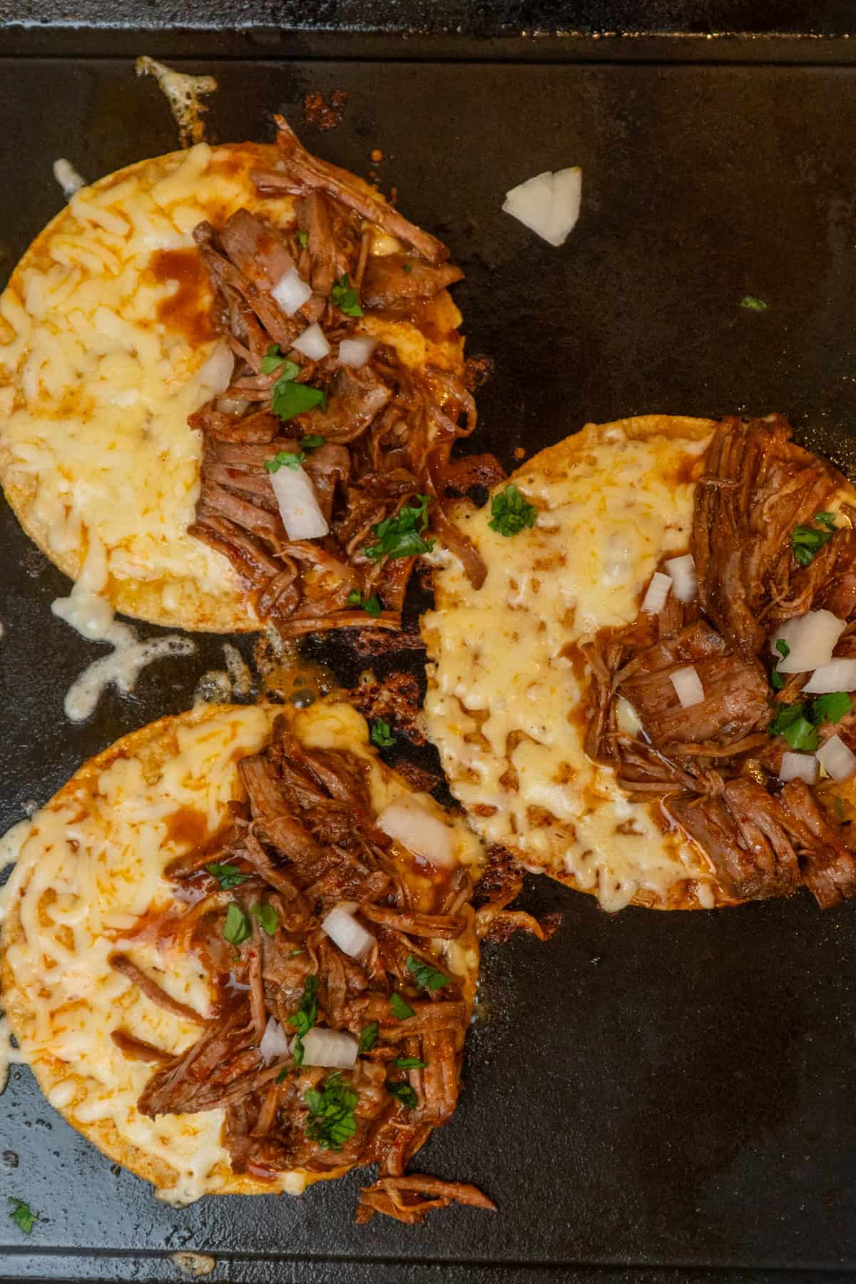 Three corn tortillas topped with cheese, chuck roast, onions and cilantro on a skillet.