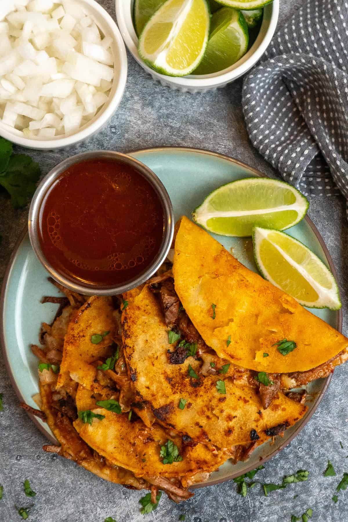 Three birria taco on a plate with limes and a bowl of birria consume.