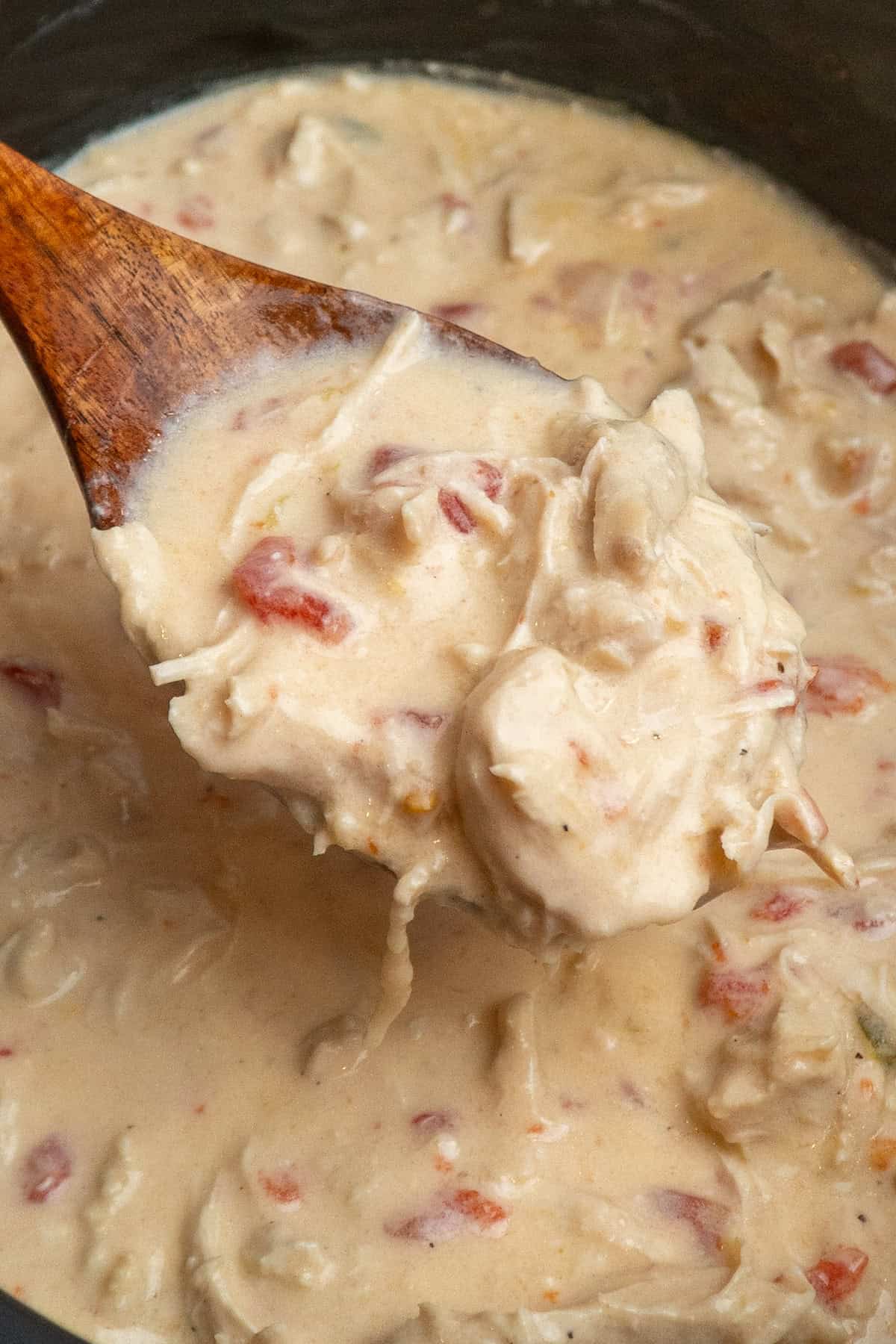 A wooden spoon holding cheesy chicken over a crock pot.