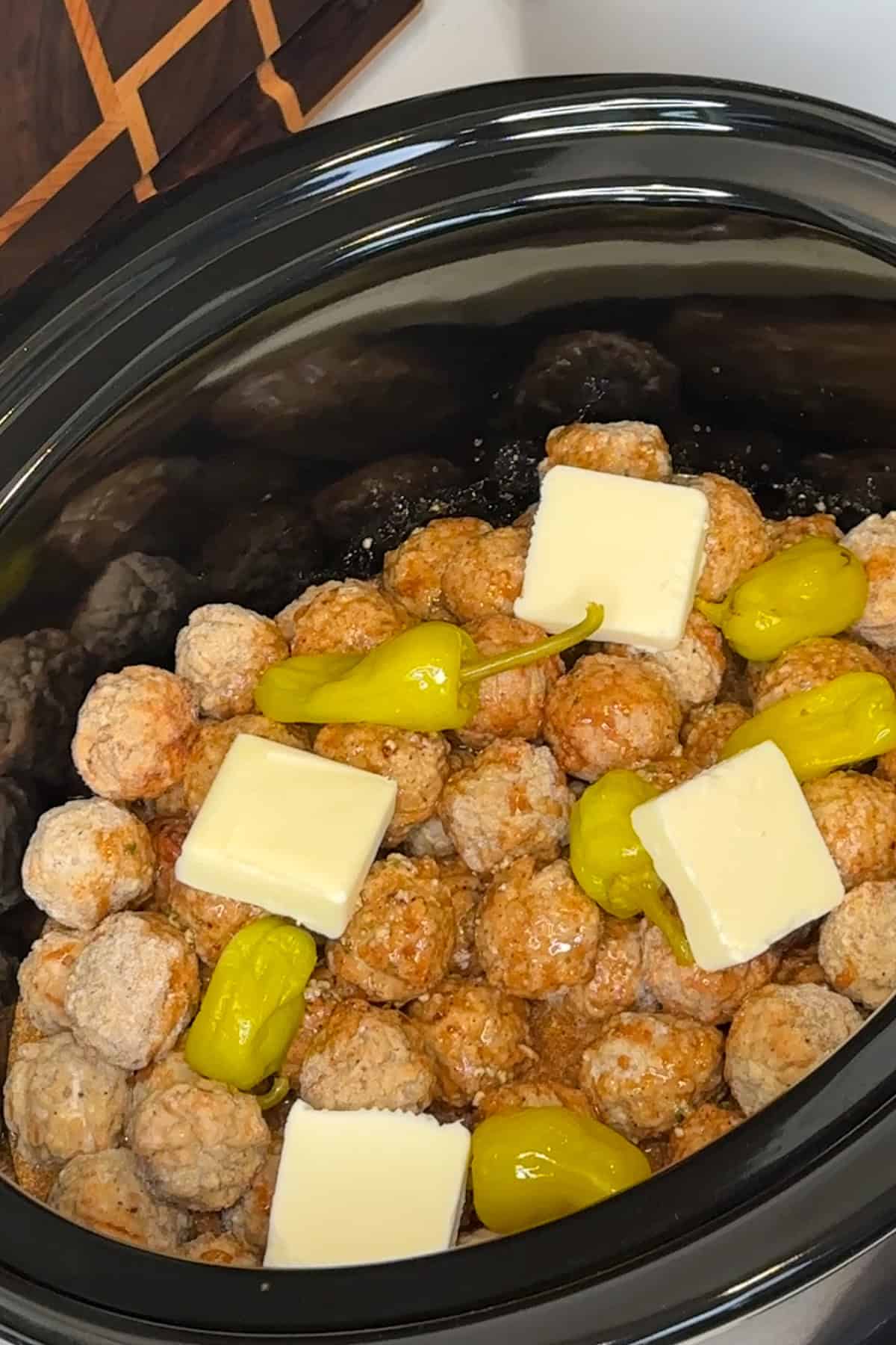 Sauce, butter, and pepperoncini peppers on top of meatballs in a crock pot.
