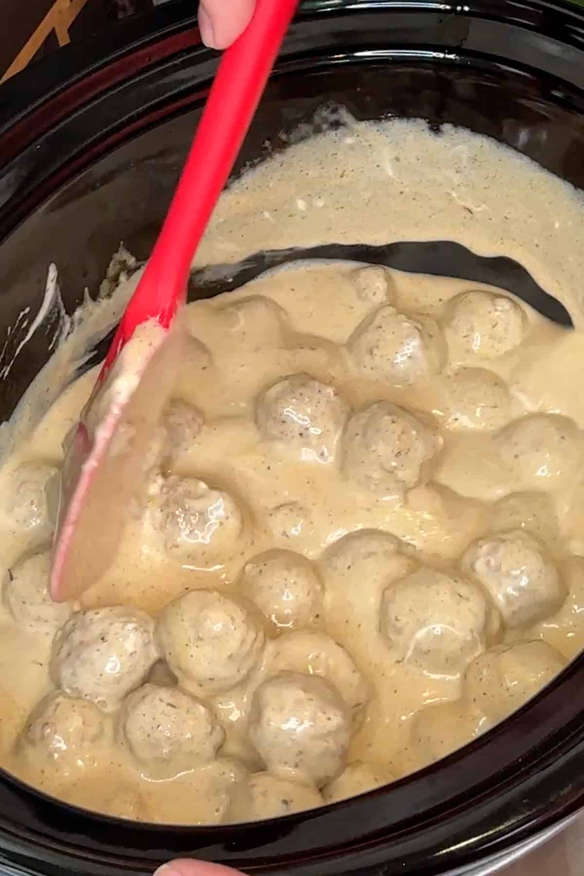 A spatula mixing together heavy cream, cornstarch, sour cream, and meatballs in a crock pot.