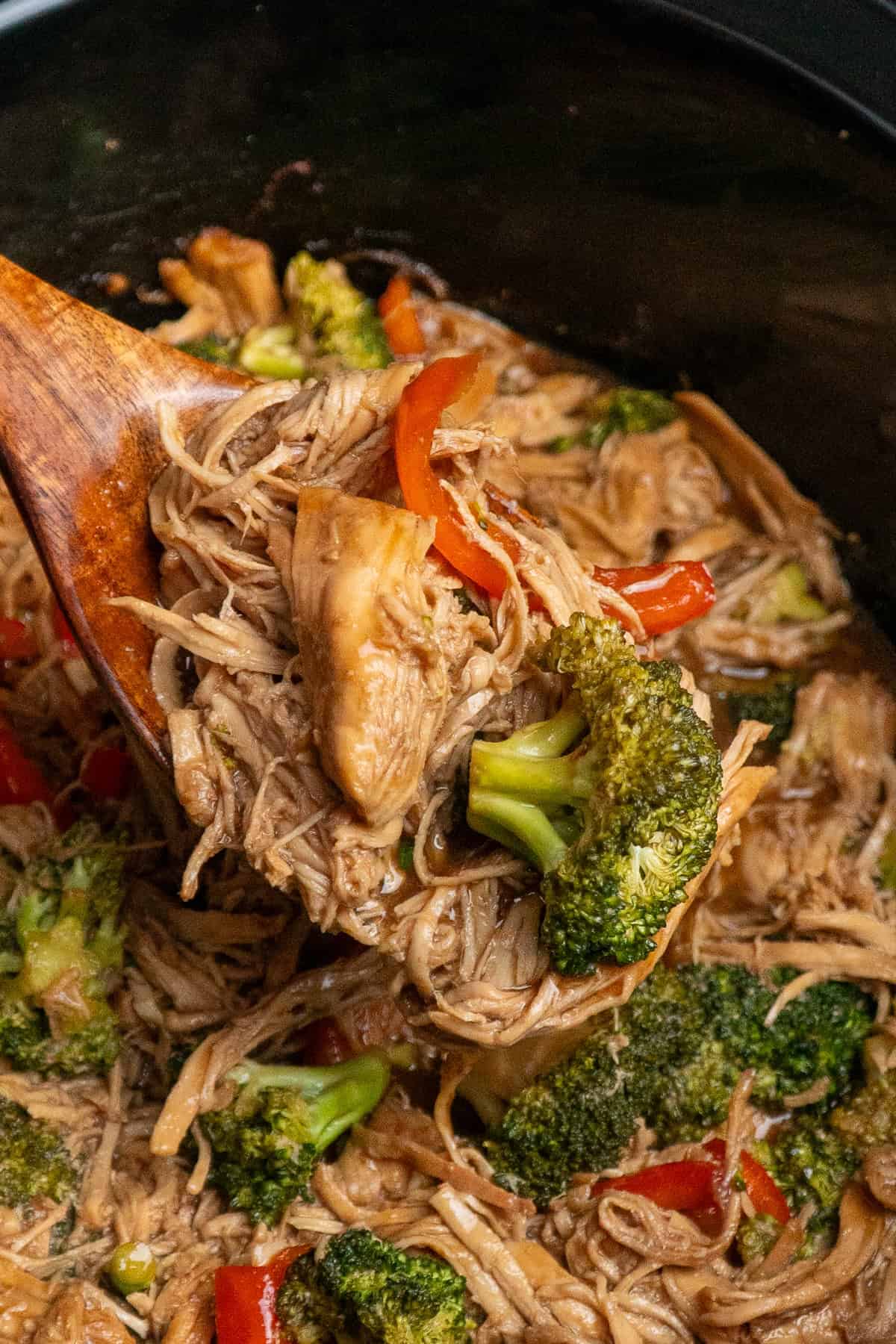 A wooden spoon holding a scoop of teriyaki chicken over a crock pot.