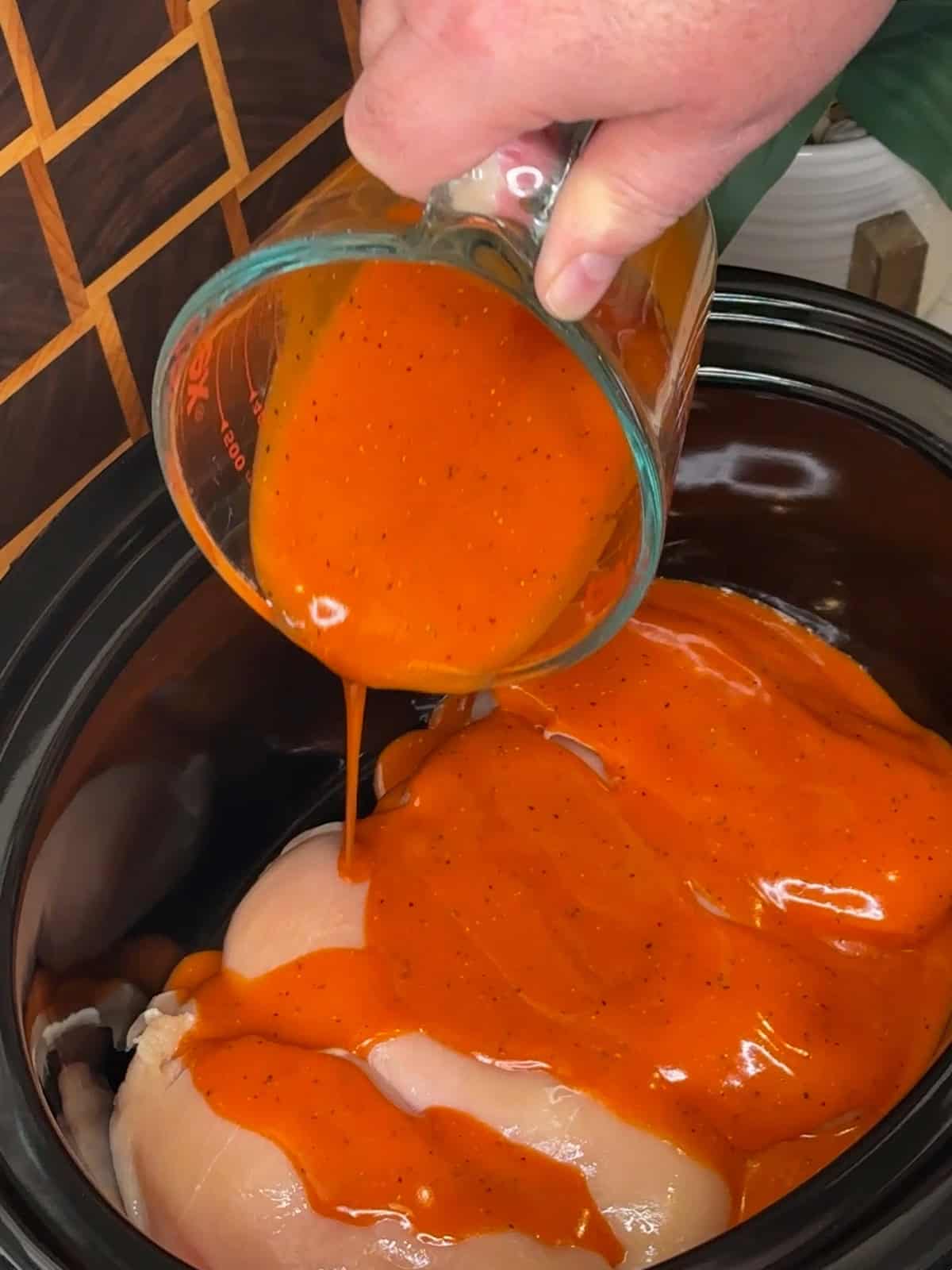 Homemade buffalo sauce being poured over chicken in a slow cooker.